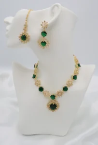 FAS jewelry set with emerald-colored stones and floral motifs, including a necklace, matching earrings, and a maang tikka in stainless steel with zircon accents.